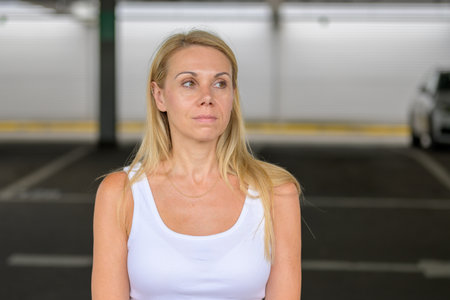 A blonde woman in a white tank top standing in a parking garage and looking to her left.の写真素材