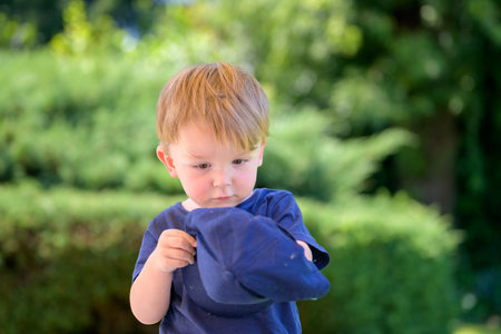 A young boy in a navy shirt looks closely at a blue cap while standing outdoors on a sunny day.の写真素材