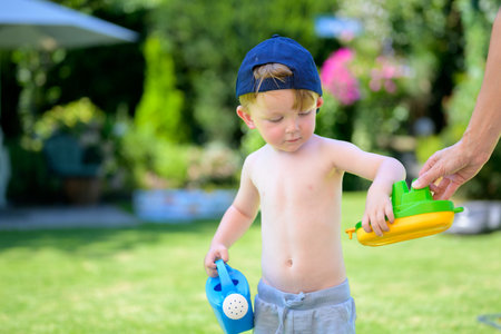 A toddler in a cap receives a colorful toy from an adult hand while holding a blue watering can.の写真素材