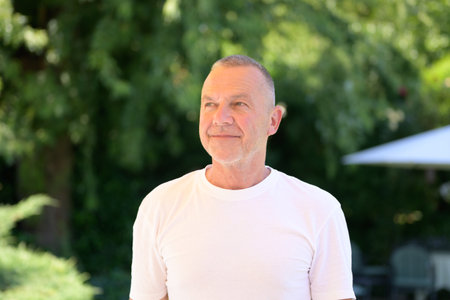 A smiling senior man in a white t-shirt looks up while standing outdoors on a bright sunny day.
