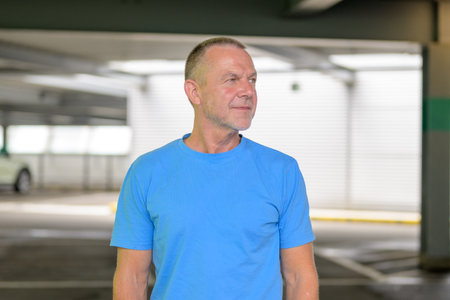 A mature man in a bright blue T-shirt stands in a modern parking structure, gazing to the side with a calm and confident demeanor.の写真素材
