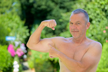 A confident older man poses shirtless outdoors, flexing his bicep and pointing at it with a smile,...