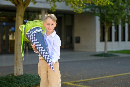 Young boy wearing a white shirt and beige pants proudly holding a decorated school cone outdoors on his first day of school.の写真素材