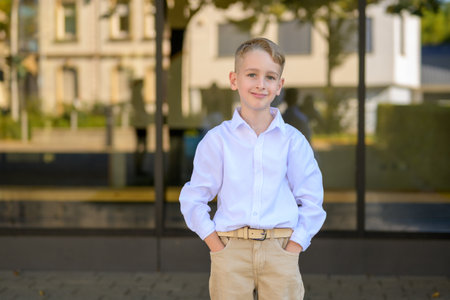 Cheerful young boy with blond hair wearing a white shirt and beige pants, smiling confidently while standing outdoors with hands in pockets.の写真素材