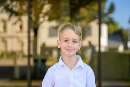 Bright outdoor portrait of a smiling young boy with blond hair and green eyes wearing a white shirt, standing confidently in warm natural sunlight.の写真素材