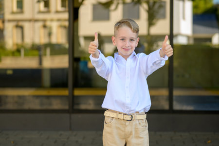 Smiling young boy with blond hair wearing a white shirt and beige pants standing outdoors, showing both thumbs up in a positive gesture.の写真素材
