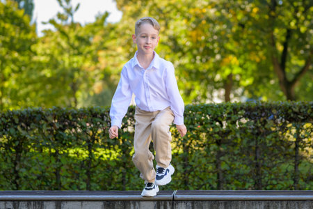 Playful young boy in beige pants and white shirt jumping down from a ledge in a sunny park with green trees in the background.の写真素材