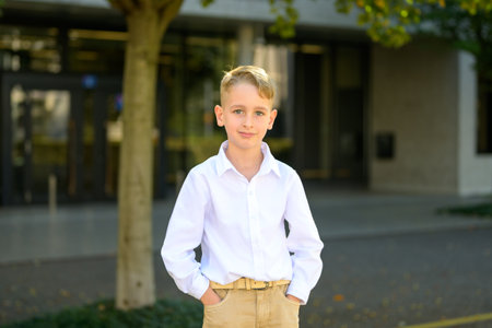 Young boy with blond hair standing confidently outdoors, wearing a white shirt and beige pants, hands in pockets, and smiling gently.の写真素材