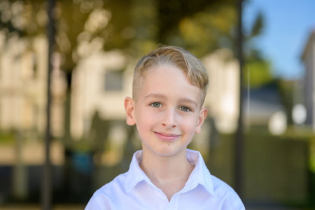 Close-up portrait of a smiling young boy with blond hair and bright eyes wearing a white shirt, standing outdoors in soft natural light.の写真素材