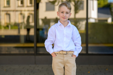 Stylish young boy with blond hair wearing a white shirt and beige pants standing outdoors with hands in pockets, looking calm and confident.の写真素材