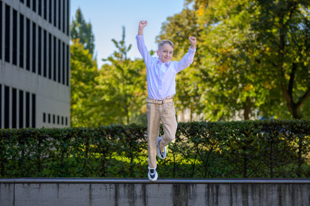 Joyful young boy in beige pants and white shirt jumping with raised arms outside on a sunny day, celebrating with energy and happiness.の写真素材