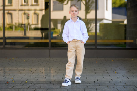 Stylish young boy with blond hair wearing a white shirt, beige pants, and sneakers standing outdoors with hands in pockets and a calm expression.の写真素材