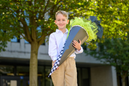 Cheerful young boy in white shirt and beige pants proudly holding a large decorated school cone outdoors on his first day of school.の写真素材