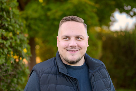 A cheerful young man with a slight smile looks to the side while standing outdoors surrounded by...