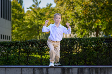 Cheerful young boy wearing a white shirt and beige pants striking a playful pose outdoors on a sunny day with green trees in the background.の写真素材