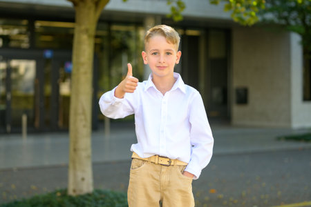 Young boy in beige pants and white shirt standing outdoors giving a confident thumbs up gesture while looking at the camera.の写真素材