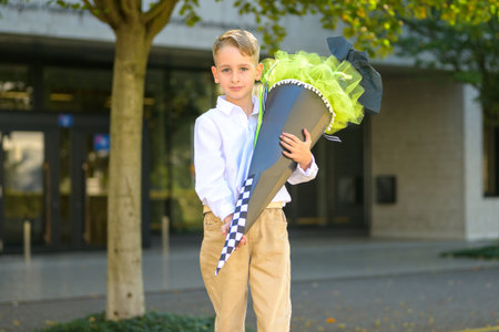 Young boy in white shirt and beige trousers holding a large decorated school cone outdoors on his first day of school.の写真素材