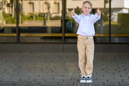 Cheerful young boy with blond hair wearing a white shirt and beige pants standing outdoors, smiling and showing both thumbs up confidently.の写真素材