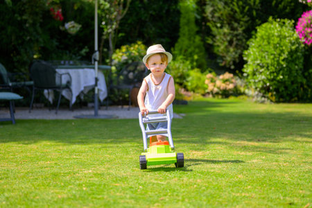 A young boy wearing a hat pushes a toy lawn mower across the grass in a sunny garden.の写真素材