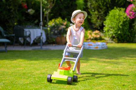 A cheerful young boy in a hat pushes a toy lawn mower across the grass on a sunny day.の写真素材