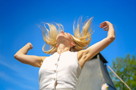 A blonde woman joyfully tosses her hair back with arms raised while enjoying a sunny day outdoors.の写真素材