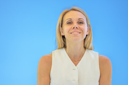 A blonde woman in a sleeveless top smiles softly while posing outdoors against a bright blue sky.の写真素材