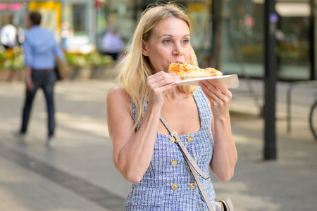 Blonde woman enjoying a fresh pizza slice while walking through the city on a bright summer day.の写真素材