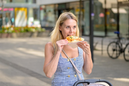 Blonde woman taking a bite of pizza outdoors while walking with a stroller on a sunny city street.の写真素材