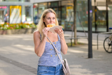 Blonde woman enjoying a hot pizza slice while standing on a sunny city street in summer.の写真素材