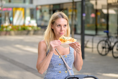 Blonde woman standing outside on a sunny day, holding a fresh pizza slice while walking with a stroller.の写真素材