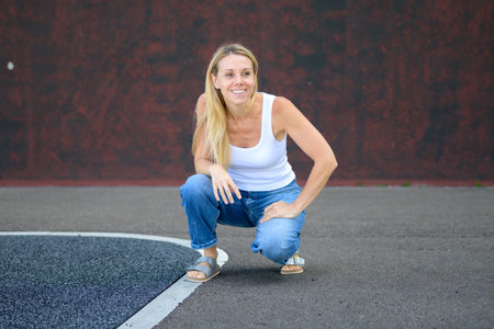 Blonde woman in casual white tank top and jeans squatting on pavement outdoors, relaxed and cheerful on a sunny day.の写真素材
