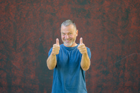 Smiling middle-aged man wearing a blue t-shirt giving double thumbs up while standing outdoors against a textured red wall.の写真素材