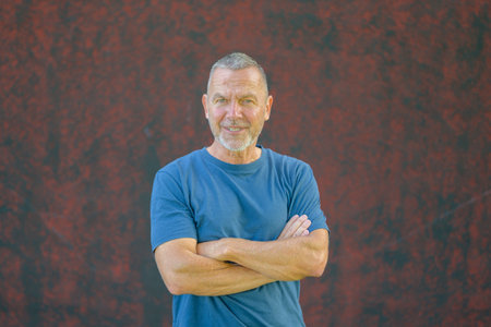 Middle-aged man with short hair and beard wearing a blue t-shirt, standing confidently with crossed arms against a textured red wall outdoors.の写真素材