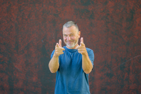 Cheerful middle-aged man in a blue t-shirt smiling and pointing forward with both hands while standing against a textured red wall outdoors.の写真素材