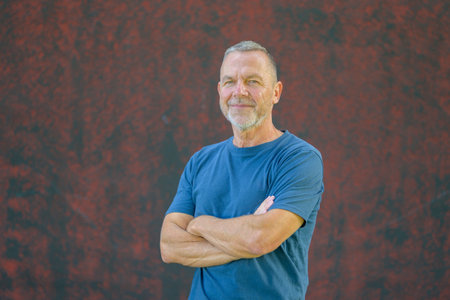 Middle-aged man with short hair and beard wearing a blue t-shirt, smiling confidently with crossed arms against a red textured wall.の写真素材