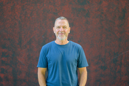 Portrait of a confident middle-aged man with short hair and beard wearing a blue t-shirt, smiling calmly against a textured red wall.の写真素材