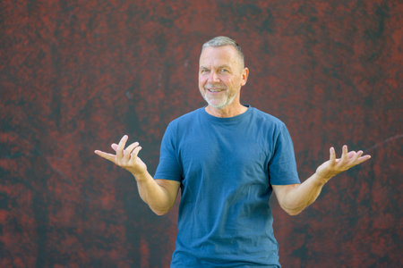 Cheerful middle-aged man in a blue t-shirt smiling and gesturing with open hands outdoors against a textured red wall, expressing positivity.の写真素材