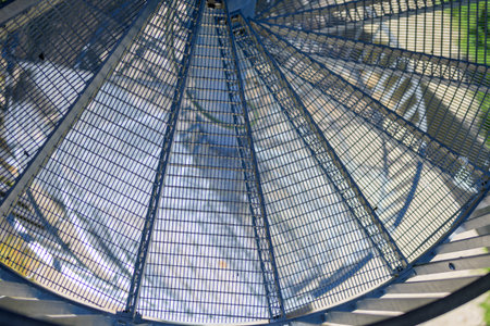 Abstract view of a blue metal spiral staircase with sunlight casting geometric shadows and reflections on the ground below.の写真素材