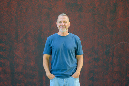 Confident middle-aged man with short hair and beard wearing a blue t-shirt and jeans, standing casually against a red textured wall with hands in pockets.の写真素材
