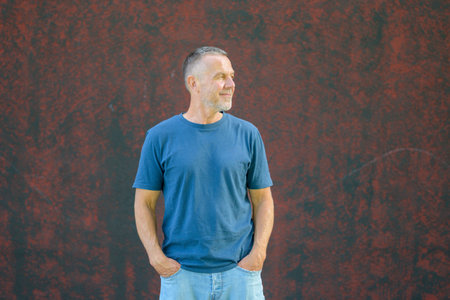 Confident middle-aged man with short hair wearing a blue t-shirt and jeans, standing outdoors against a textured red wall with hands in pockets.の写真素材
