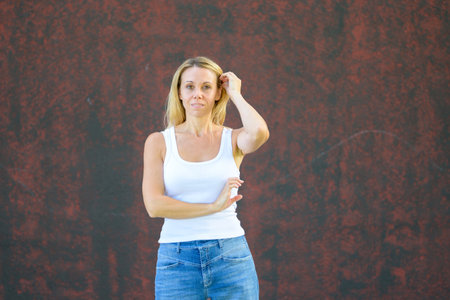 Blonde woman in casual white tank top and jeans standing outdoors against a textured red wall, posing naturally with calm confidence.の写真素材
