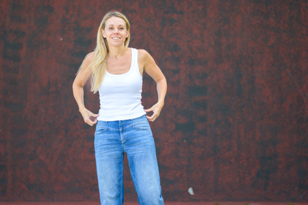 Cheerful blonde woman in casual white tank top and blue jeans standing outdoors against a textured red wall, looking relaxed and confident.の写真素材