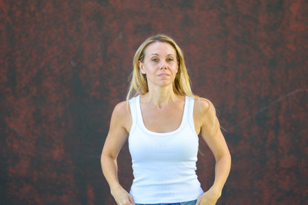 Blonde woman in casual white tank top and jeans standing outdoors against a textured red wall, looking confident and composed.の写真素材