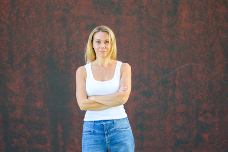 Blonde woman in casual white tank top and jeans standing with crossed arms against a textured red wall, looking calm and self-assured.の写真素材