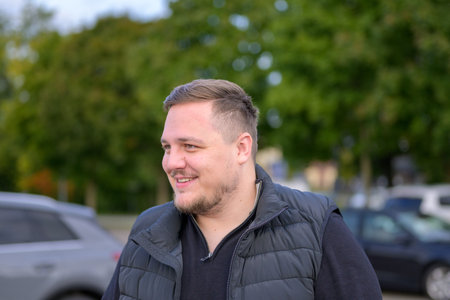 Portrait of a smiling young man wearing a black jacket outdoors on an autumn day with green trees in...