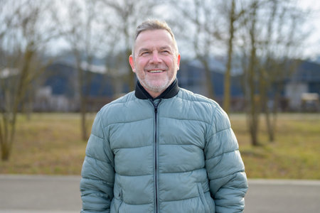 Smiling mature man wearing a padded jacket while standing outdoors in a park, looking forward with a...