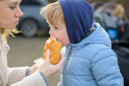 A toddler wearing a warm jacket is eating bread while being gently assisted by an adult outdoors.