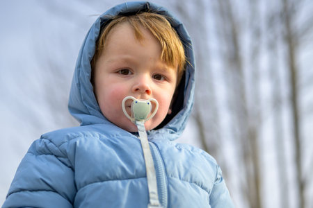 Close up portrait of a toddler wearing a blue winter jacket and pacifier outdoors, showing a calm...