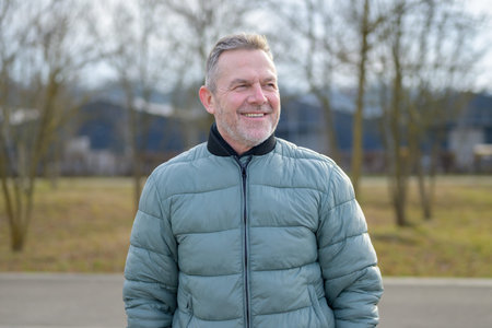 Smiling mature man wearing a padded jacket while standing outdoors in a park, looking to the side...