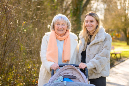 Happy mother and grandmother walking together with baby stroller in park. Authentic...